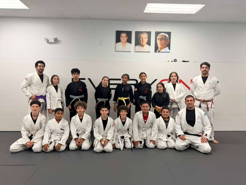 A full kids and teens jiu-jitsu class posing in gis with their coaches under the Everyone Jiu Jitsu wall logo